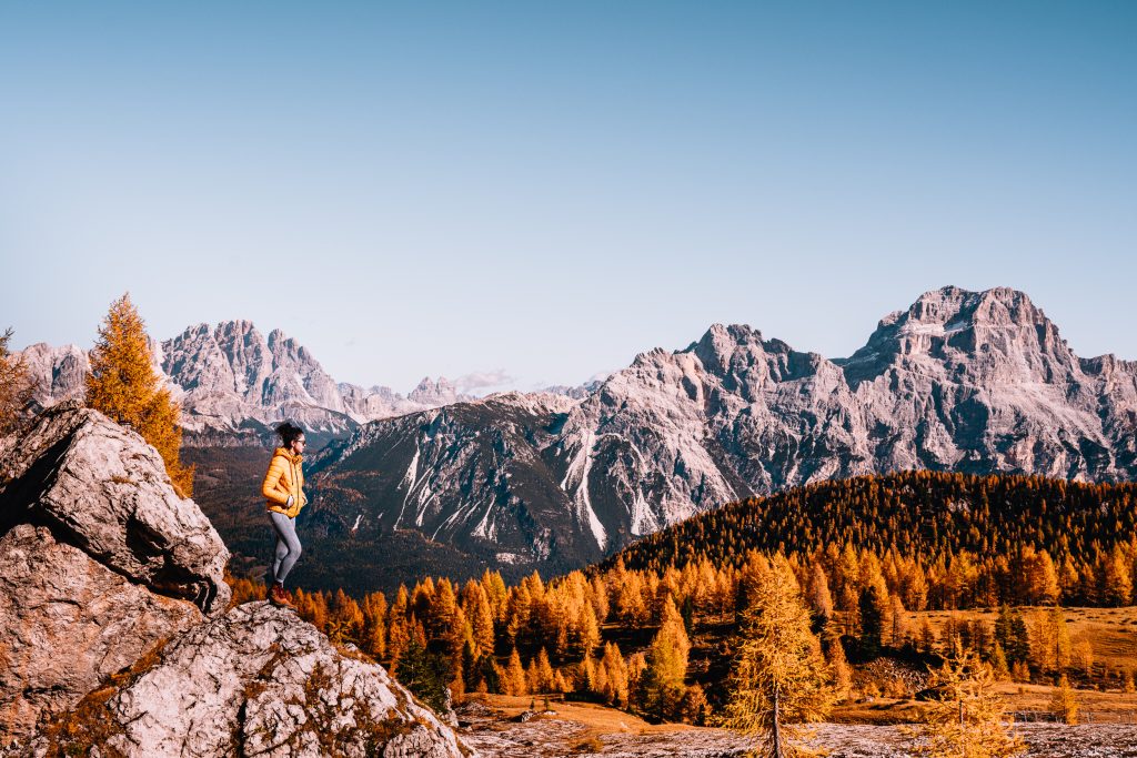 A woman in an orange puffa jacket stands on an outcrop of rock looking out over bright orange larch trees and mountains in the distance on the Croda da Lago hike, one of my favourites to put on a Dolomites itinerary
