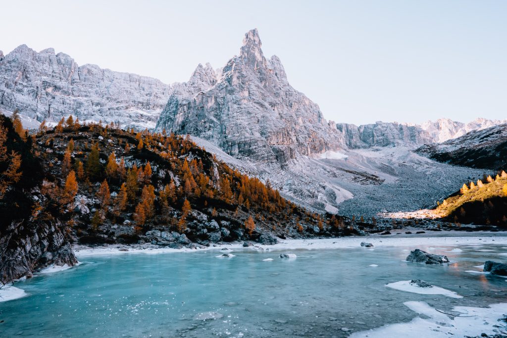 The turquoise water of frozen Lago di Sorapis in the foreground with golden larch trees and a mountain peak in the distance