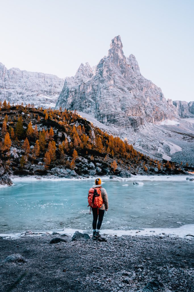 a woman with an orange backpack stands in front of the frozen turquoise water of lake sorapis